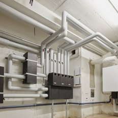 Ein technischer Raum mit zahlreichen weißen Rohren, die an der Wand verlaufen, und einer Heizungsanlage in der Ecke.