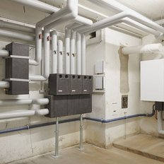 Ein technischer Raum mit zahlreichen Rohren und Ventilen, die an einer Wand montiert sind, zeigt eine komplexe Heizungsinstallation.