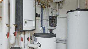 Ein Heizungsraum mit verschiedenen Rohren, einem großen weißen Warmwasserspeicher und einem Steuergerät an der Wand.
