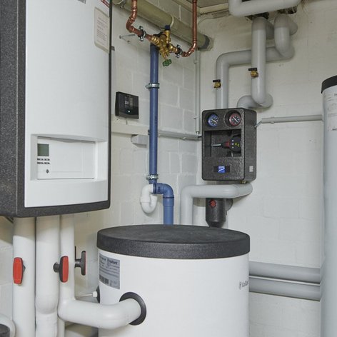 Ein Heizungsraum mit verschiedenen Rohren, einem großen weißen Warmwasserspeicher und einem Steuergerät an der Wand.