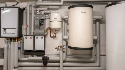 Ein technischer Raum mit Heizungsanlagen, Rohren und einem großen weißen Warmwasserspeicher an der Wand.