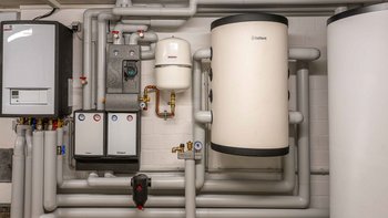 Ein technischer Raum mit Heizungsanlagen, Rohren und einem großen weißen Warmwasserspeicher an der Wand.