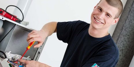 Ein junger Techniker arbeitet mit einem Schraubendreher an einem offenen Gehäuse, lächelt dabei freundlich in die Kamera.