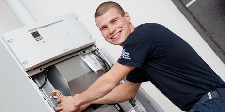 Ein junger Techniker lächelt, während er an einer Heizungsanlage arbeitet, umgeben von modernen Geräten und Kabeln.