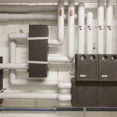 Die Installation zeigt mehrere Rohre und Kastenstrukturen, die in einem technischen Raum für Heizungsanlagen angeordnet sind.