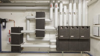 Die Installation zeigt mehrere Rohre und Kastenstrukturen, die in einem technischen Raum für Heizungsanlagen angeordnet sind.