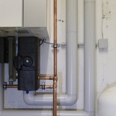 Eine Heizungsanlage mit verschiedenen Rohren und einem großen weißen Wassertank ist an einer Wand installiert.