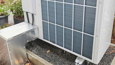 Eine moderne Klimaanlage steht auf einem stabilen Fundament aus Kies, umgeben von grünen Pflanzen und Blumentöpfen.