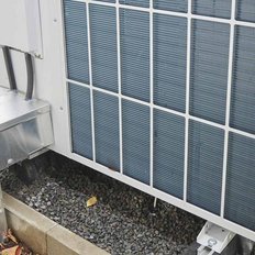 Eine moderne Klimaanlage steht auf einem stabilen Fundament aus Kies, umgeben von grünen Pflanzen und Blumentöpfen.