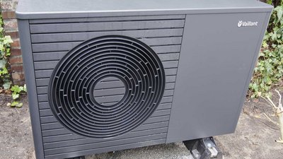 Eine moderne Wärmepumpe in einem Garten, umgeben von grünen Pflanzen und einem Ziegelmauerhintergrund.