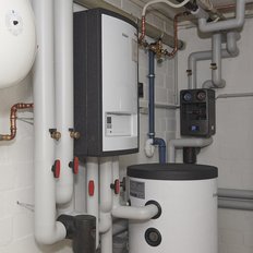 Ein Heizungsraum mit verschiedenen Rohren, einem großen weißen Warmwasserspeicher und einem modernen Heizgerät an der Wand.