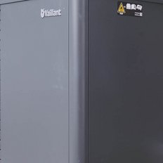 Eine moderne Wärmepumpe steht an einer Ziegelmauer, umgeben von grünen Pflanzen, die für Energieeffizienz sorgt.