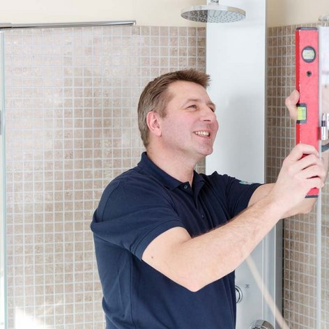 Ein lächelnder Mann arbeitet an der Installation einer Duschabtrennung und verwendet eine Wasserwaage zur genauen Ausrichtung.