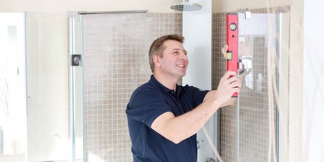 Ein lächelnder Mann arbeitet an der Installation einer Duschabtrennung und verwendet eine Wasserwaage zur genauen Ausrichtung.