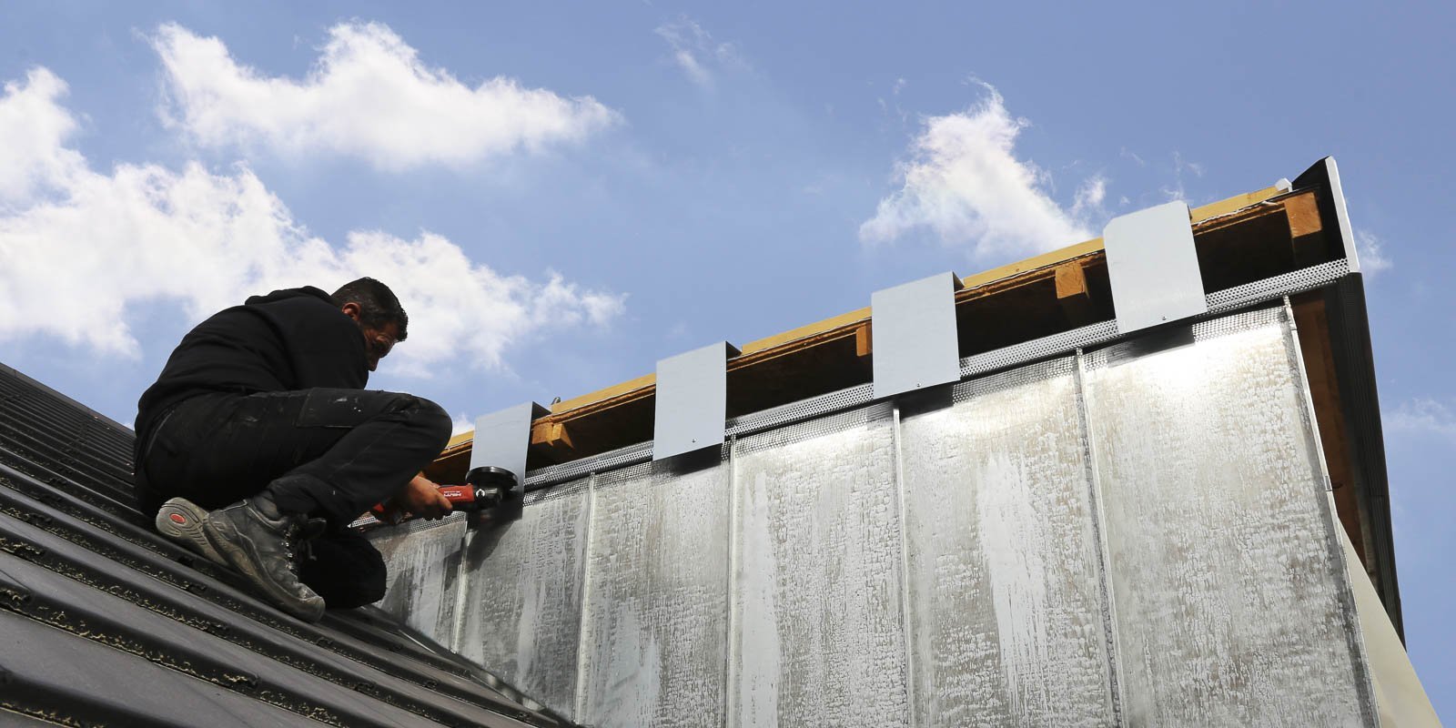 Ein Handwerker arbeitet konzentriert auf einem Dach und befestigt Metallplatten mit einem Werkzeug, während der Himmel blau ist.